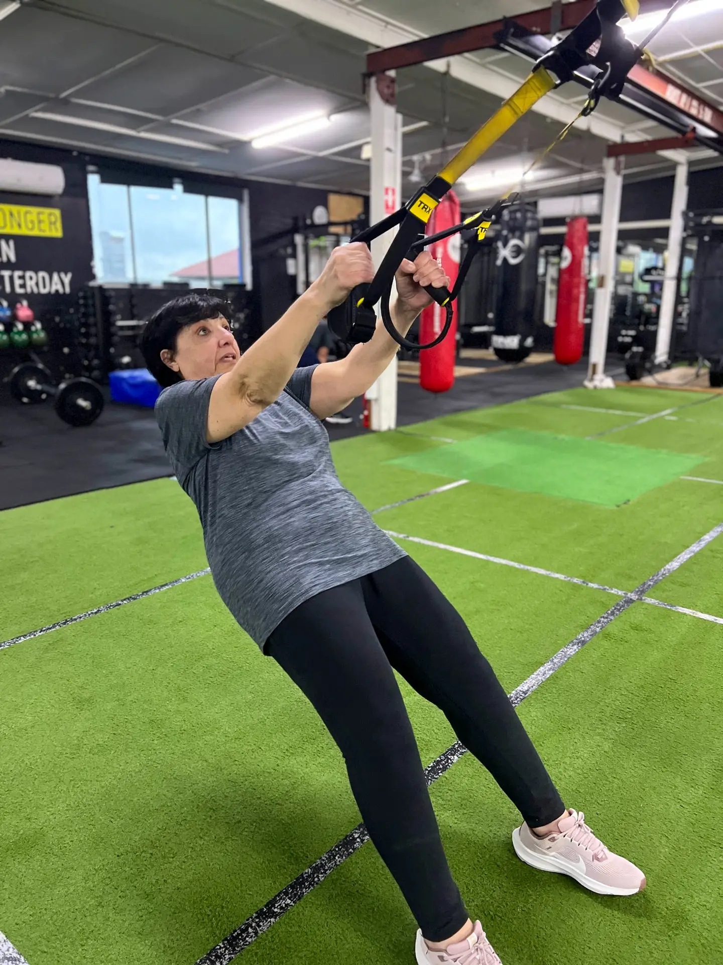 An older woman performs a suspended bodyweight row using TRX straps on the turf floor at Catalyst Training Co. gym in Randwick.