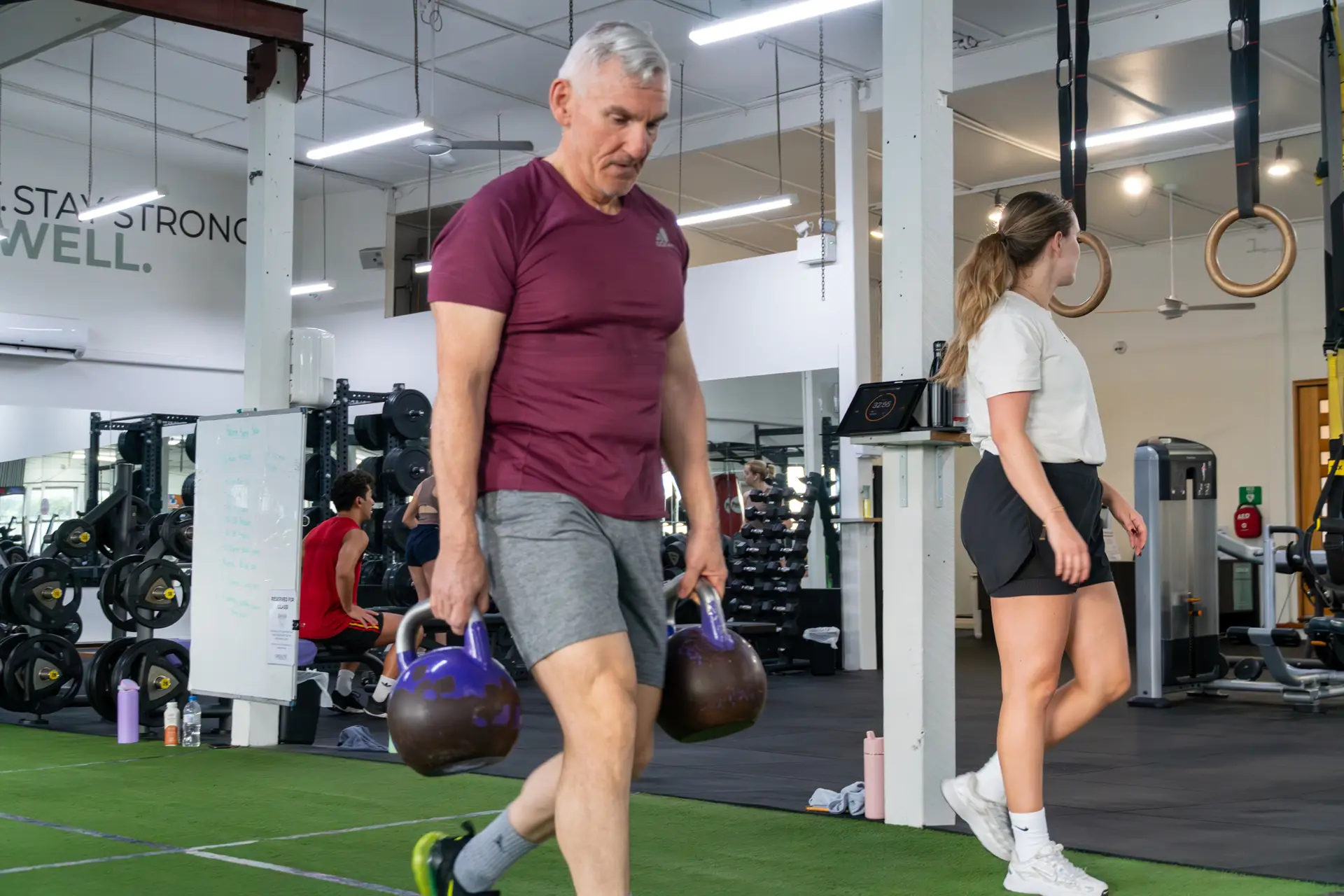 A Catalyst member carrying two heavy kettlebells in a farmer's walk on the gym floor in Randwick