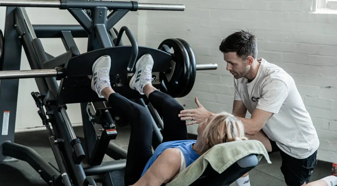 A coach training an older woman as she performs a leg press on a weight machine at a functional fitness gym in Randwick.