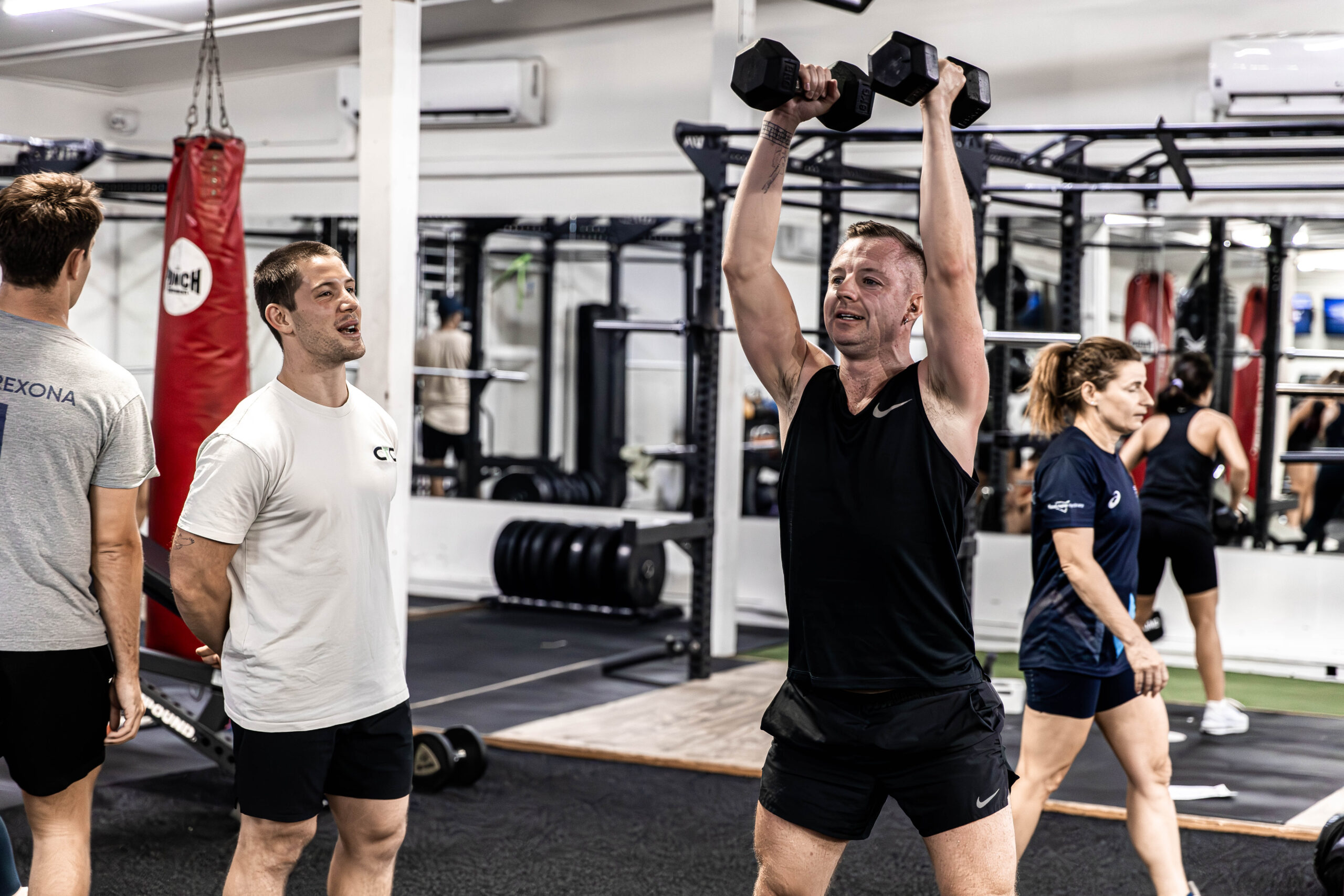 A member pressing dumbbells overhead while a Catalyst coach watches, on the gym floor in Randwick